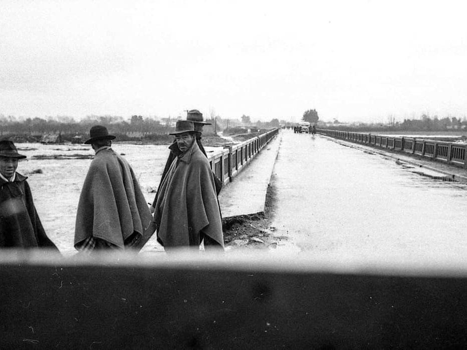 Fotografía monocromática, en primer plano se aprecian cuatro hombres vestidos con ponchos y sombreros. En segundo plano, se divisa un rio sobrepasado por un puente que al final de la toma aparece un microbús rodeado por personas.