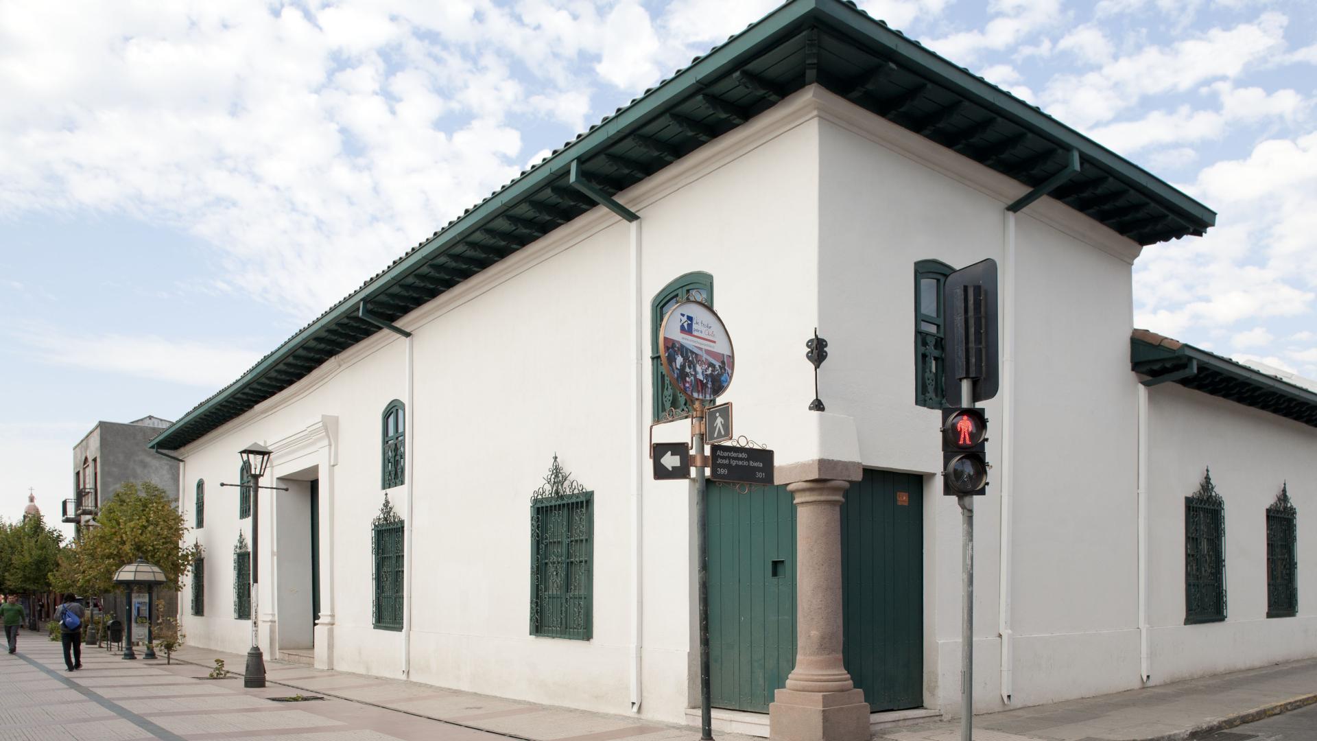 Casa de dos pisos del periodo colonial en tono blanco y detalles verdes. En su esquina se muestra en su esquina un pilar de piedra.