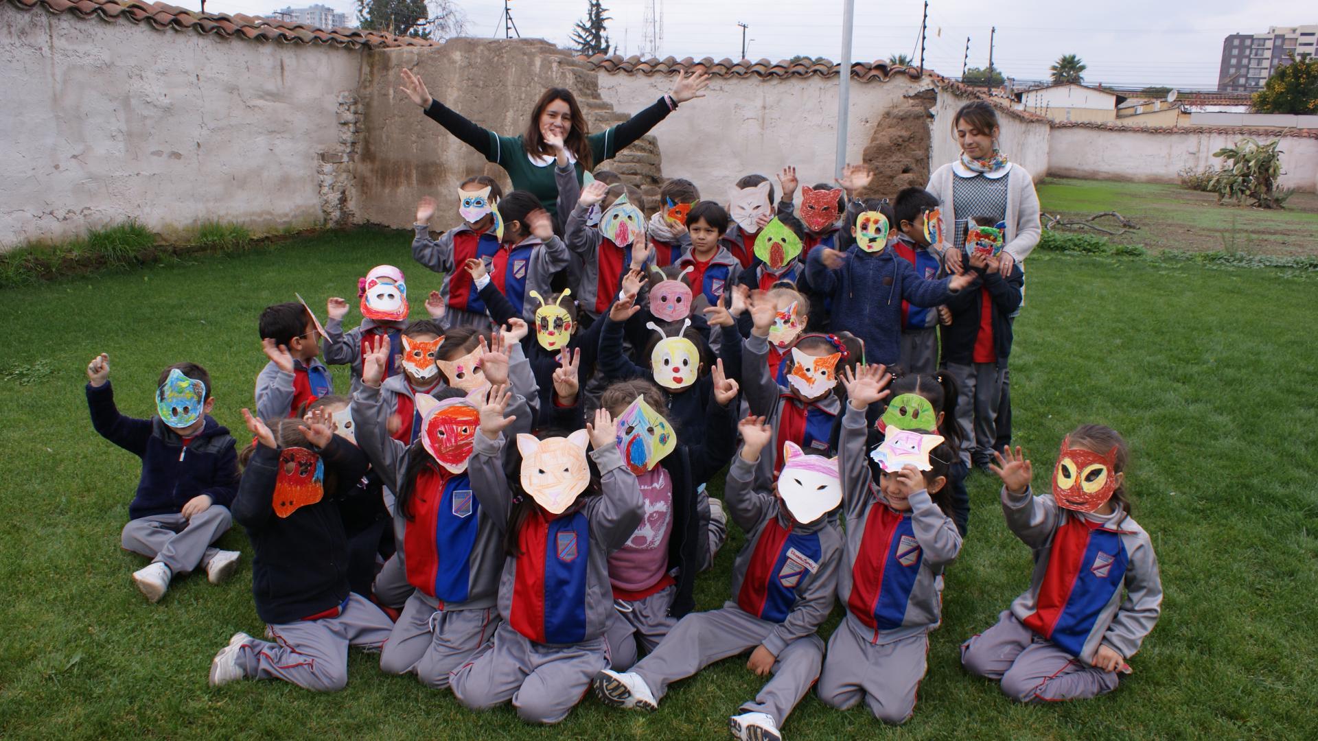 Grupos escolar de niñas y niños junto a educadoras de parvulo saludando a la cámara. Los niños y niñas llevan puesta máscaras de papel que representan fauna en riesgo presentes en la región tales como el loro tricahue, zorro cumpeo, ranita de darwin, entre otros.