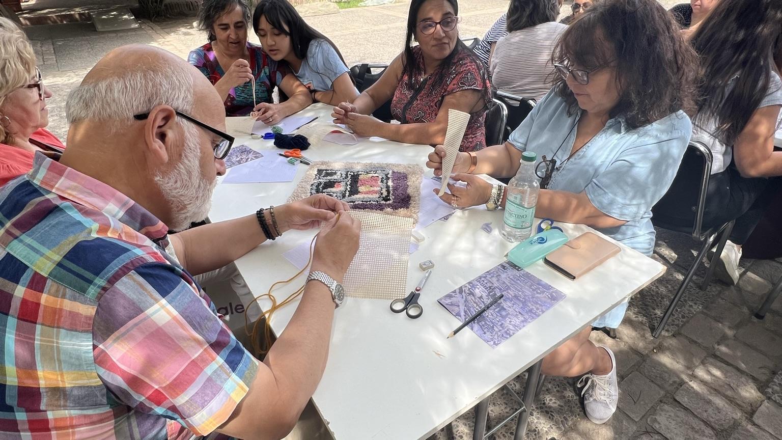Se aprecian personas adultas sentadas alrededor de una mesa, ellos se encuentran concentrados haciendo un tejido. De fondo, aparecen arbustos y la casa colonial que alberga el museo. 