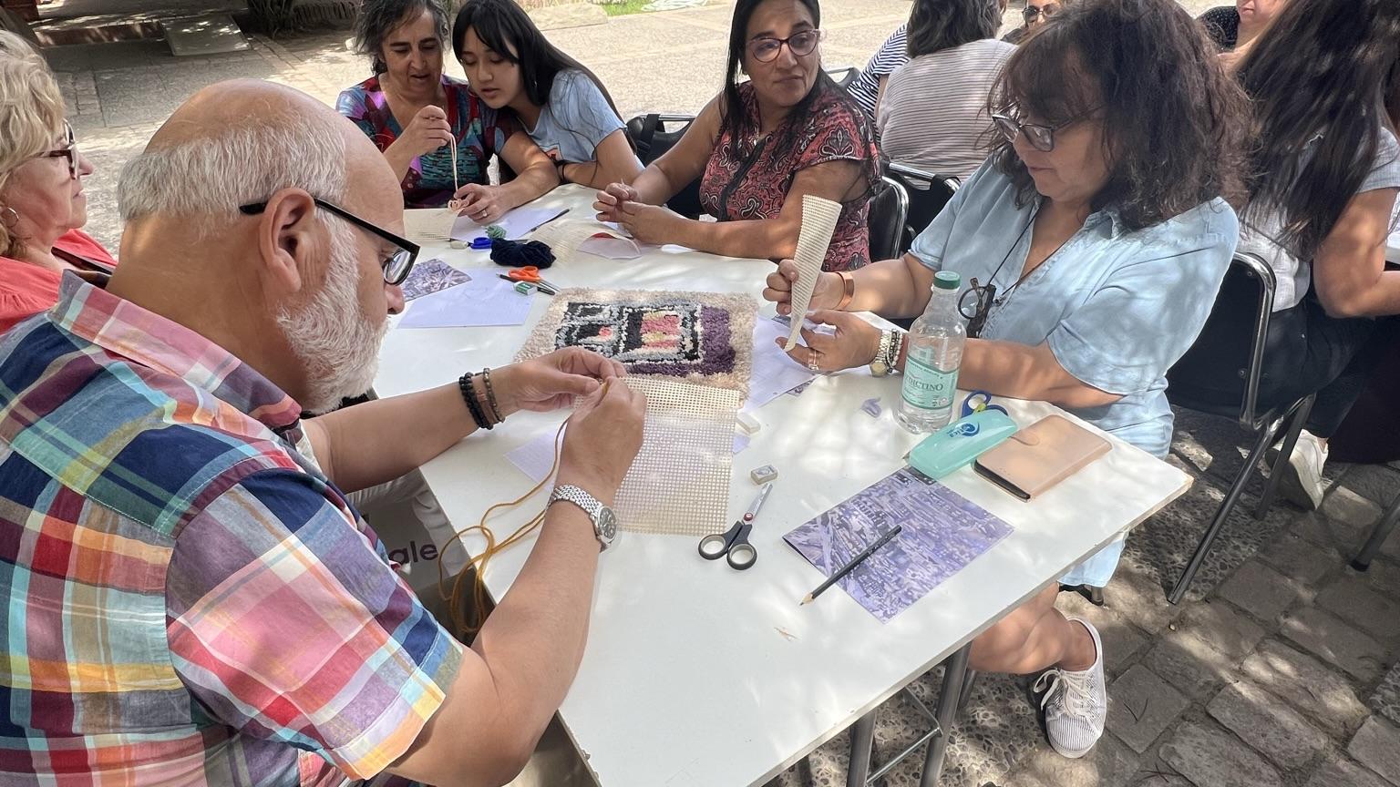 Se aprecian personas adultas sentadas alrededor de una mesa, ellos se encuentran concentrados haciendo un tejido. De fondo, aparecen arbustos y la casa colonial que alberga el museo. 