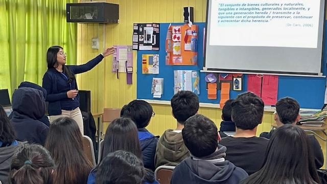 Fotografía de charla, en primer plano distintos estudiantes sentados, al fondo de la imagen se aprecia una mujer señalando un panel que tiene escrita la frase “El Patrimonio es…”