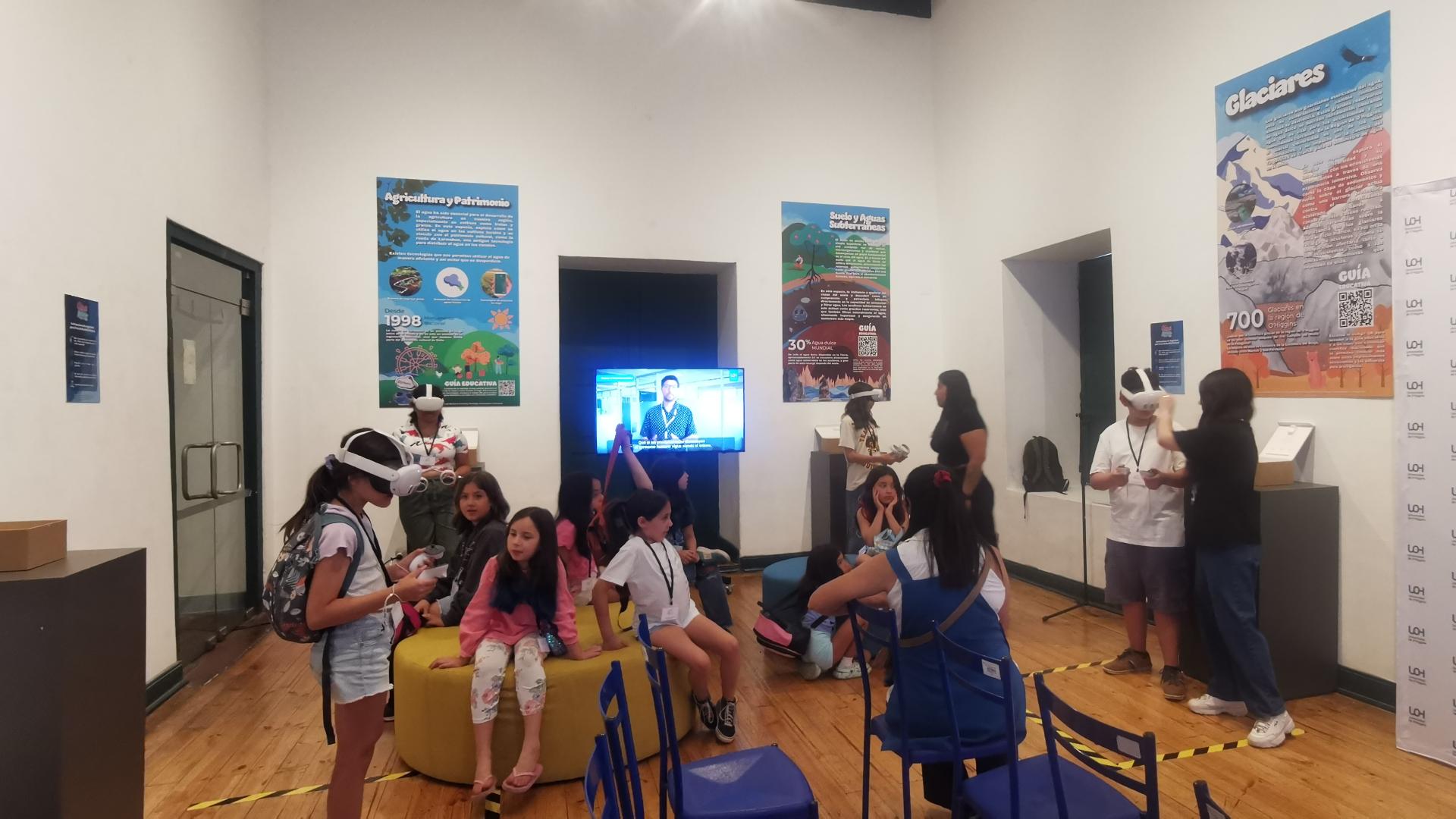 Niñas y niños de pie y sentados en un salon. Una niña con lentes VR en la cabez y las otras niñas la observan.