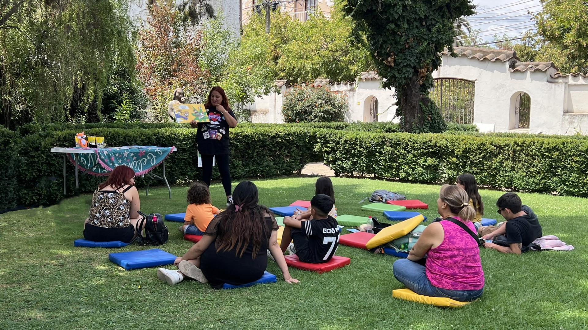 Fotografía panorámica del patio del museo, en el centro hay un grupo de niños y adultos sentados en cojines de colores dispuestos en el césped. De pie, una mujer que es la mediadora de la actividad, ella señala un libro abierto que tiene en su mano. En el fondo de la imagen algunos arboles altos. 