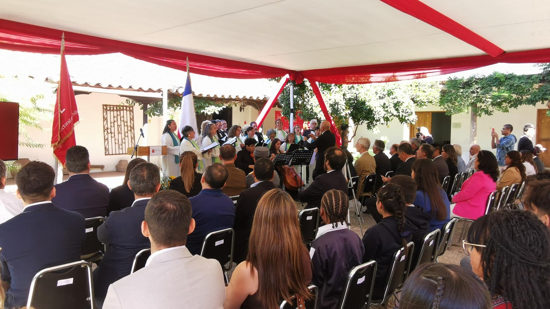 personas sentadas bajo un carpa de color blanco, al fondo personas en un coro de pie