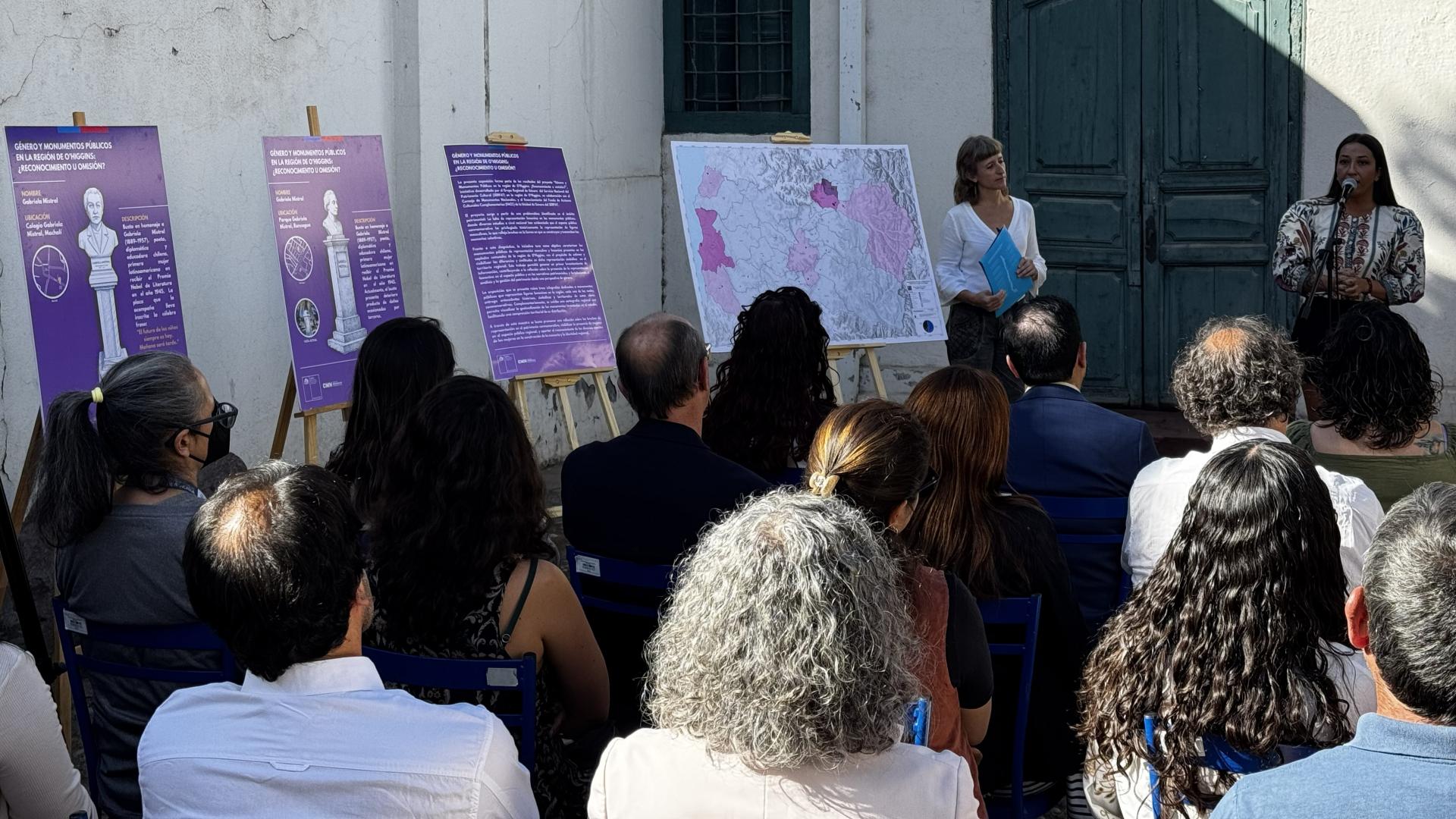 En en espacio exterior de un edificio con muros blancos y una puerta de madera, Dos mujeres están de pie, una de ellas habla con un micófono a las personas que escuchan sentadas