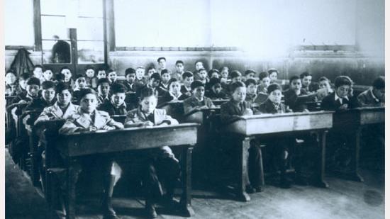 Fotografía monocromatica de una sala de clases en el Instituto O'Higgins de Rancagua. En ella se aprecia un grupo de niños sentados en sus pupitres dobles, la sala está distribuida en tres filas, en el fondo se observa una pared con una ventana de gran tamaño que aparentemente permite mirar hacia otra sala de clases.  