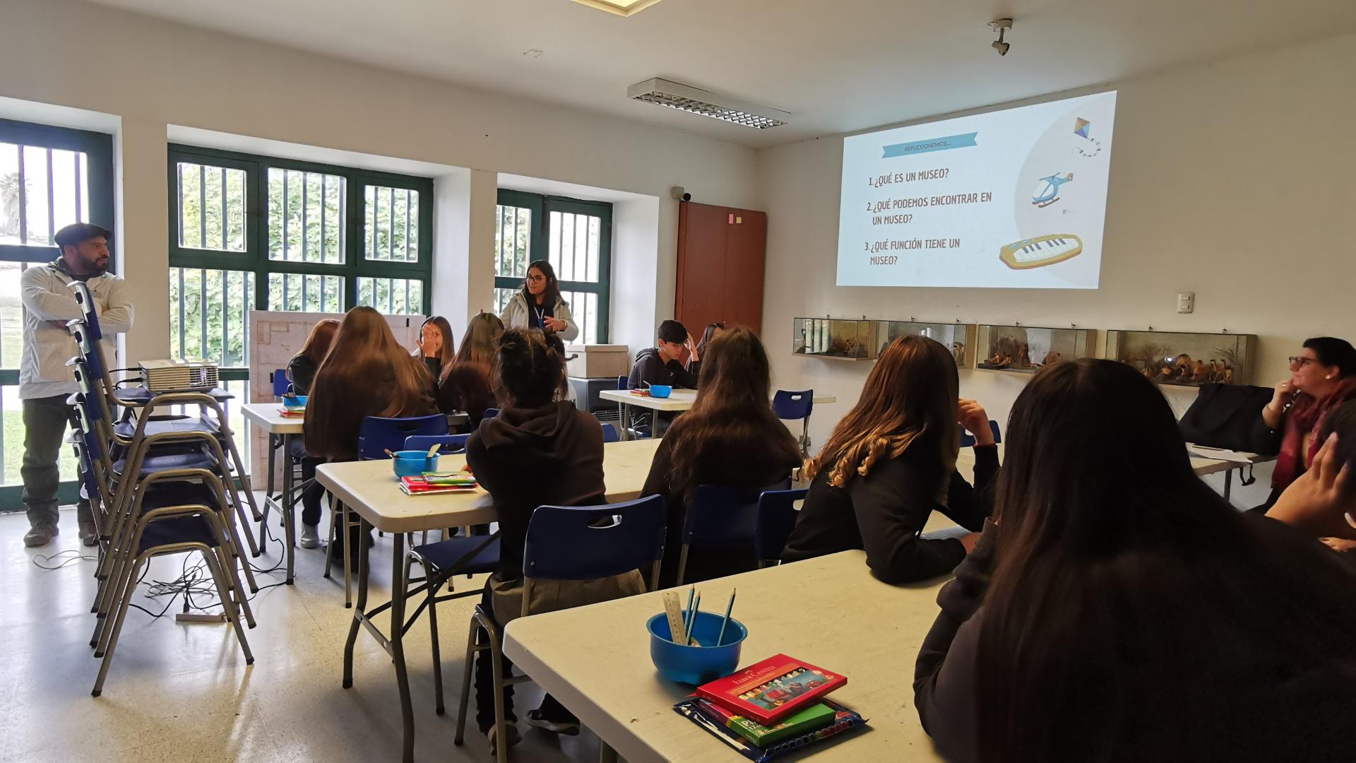 Estudiantes sentados en grupos en un salón, observan a una mujer adulta que muestra una presentación en un proyector
