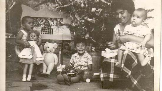 Fotografía en escala de grises en que se aprecian una niña, un niño y un bebe en los brazos de un adulto. LA escena se contextualiza en un arbol de navidad y los niños tienen en sus manos juguetes como una muñeca y un peluche. 