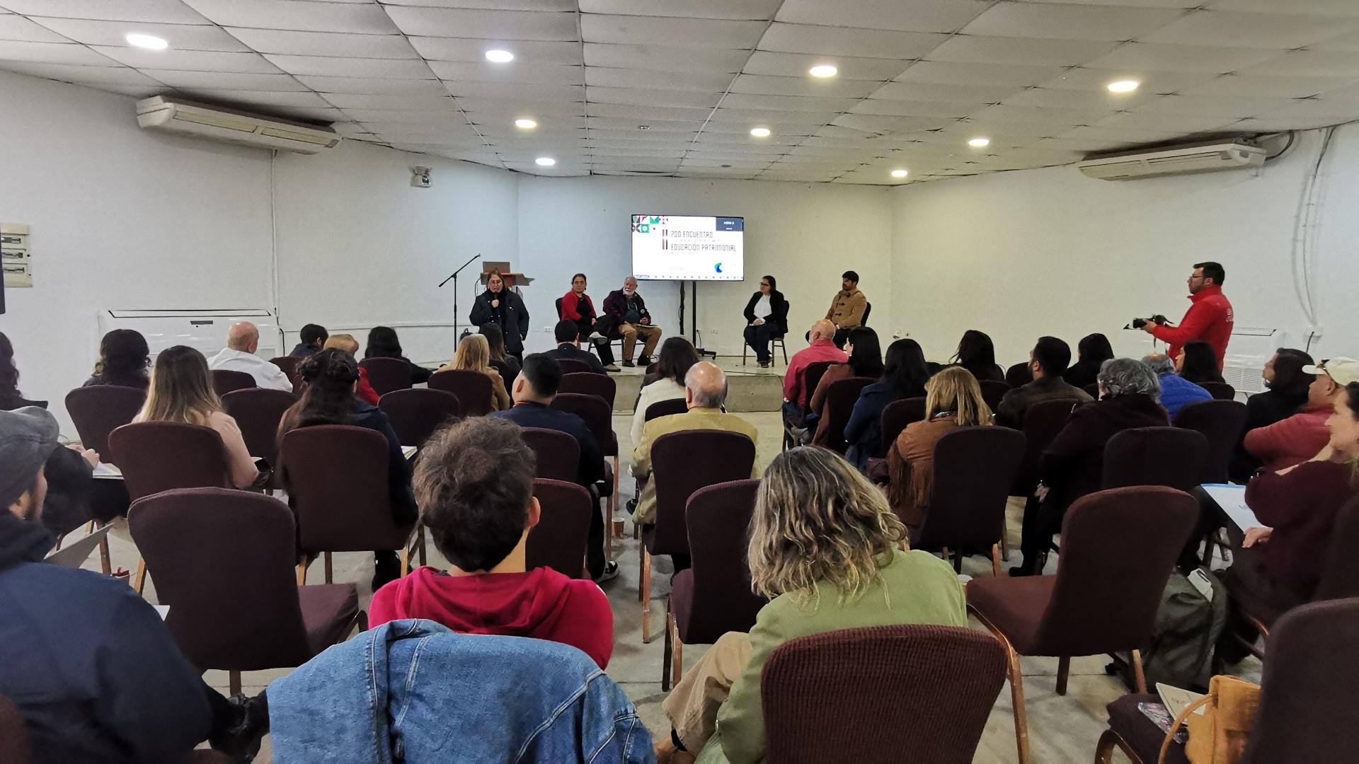personas sentadas en un salón escuchan a los expositores que presentan en el escenario