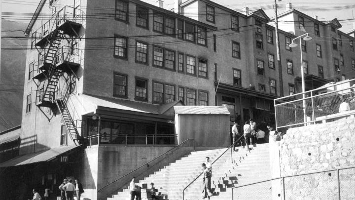 Fotografía monocromática en que se observan en primer plano personas usando la escalera central de Sewell, en segundo plano tres edificios denominados camarotes, lugar en que pernoctaban los mineros. 