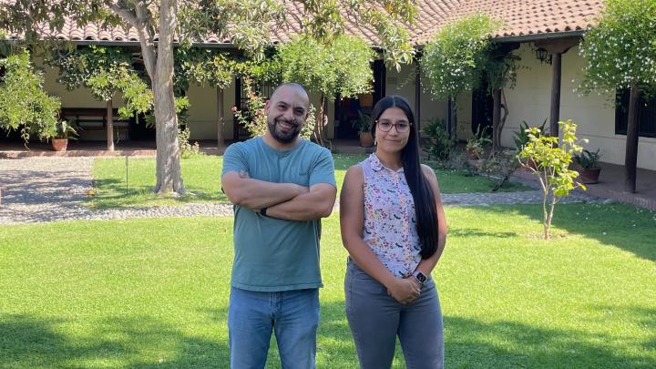 un hombre y una mujer sonríen y posan para una fotografía, detras de ellos se observa un dia soleado, pasto, arboles, arbustos y una casa colonial con tejas.