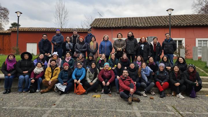 personas sentadas y de pie en el frontis de un edicicio colonial rojo, posan para una fotografía con actitud alegre
