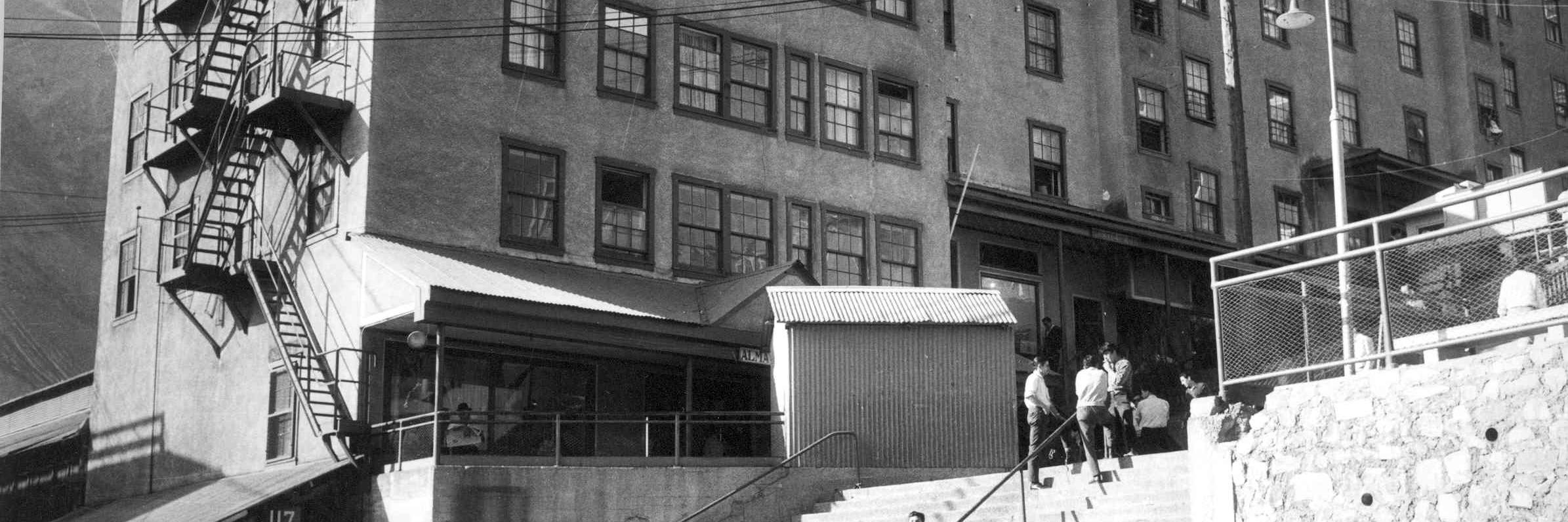 Fotografía monocromática en que se observan en primer plano personas usando la escalera central de Sewell, en segundo plano tres edificios denominados camarotes, lugar en que pernoctaban los mineros. 