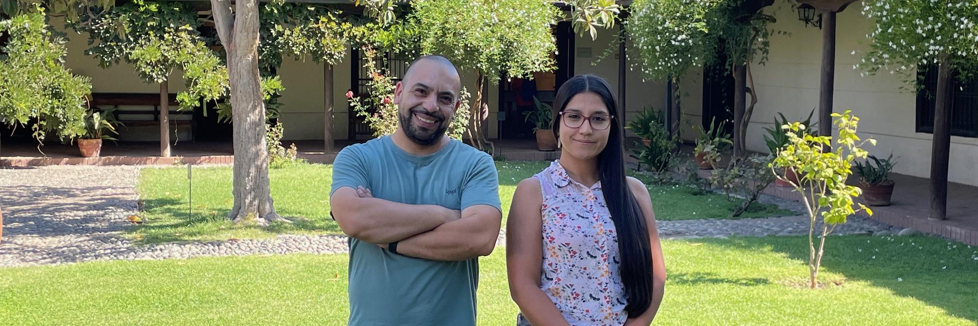 un hombre y una mujer sonríen y posan para una fotografía, detras de ellos se observa un dia soleado, pasto, arboles, arbustos y una casa colonial con tejas.