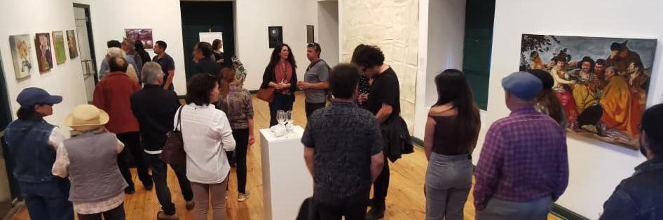 En un salón de paredes blancas y piso de madera mujeres y hombres conversan entre sí. en ls paredes se observan cuadros y tambien una escultura en un pedestal.