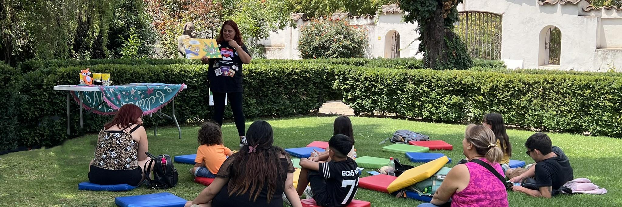 Fotografía panorámica del patio del museo, en el centro hay un grupo de niños y adultos sentados en cojines de colores dispuestos en el césped. De pie, una mujer que es la mediadora de la actividad, ella señala un libro abierto que tiene en su mano. En el fondo de la imagen algunos arboles altos. 