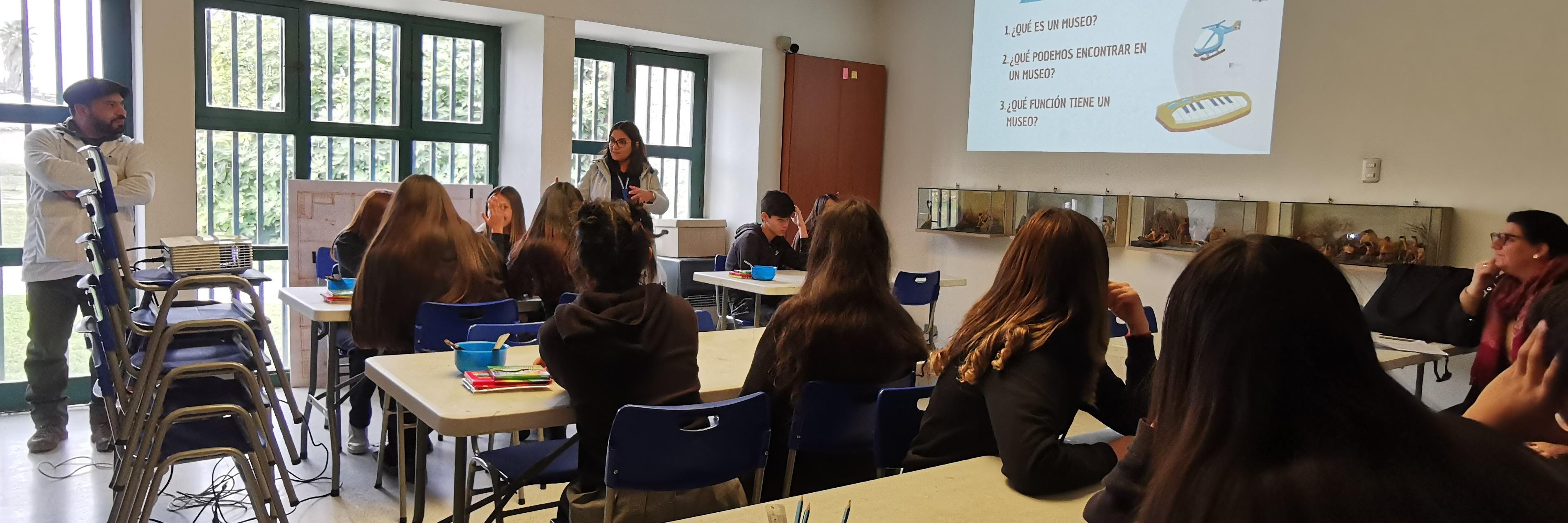 Estudiantes sentados en grupos en un salón, observan a una mujer adulta que muestra una presentación en un proyector