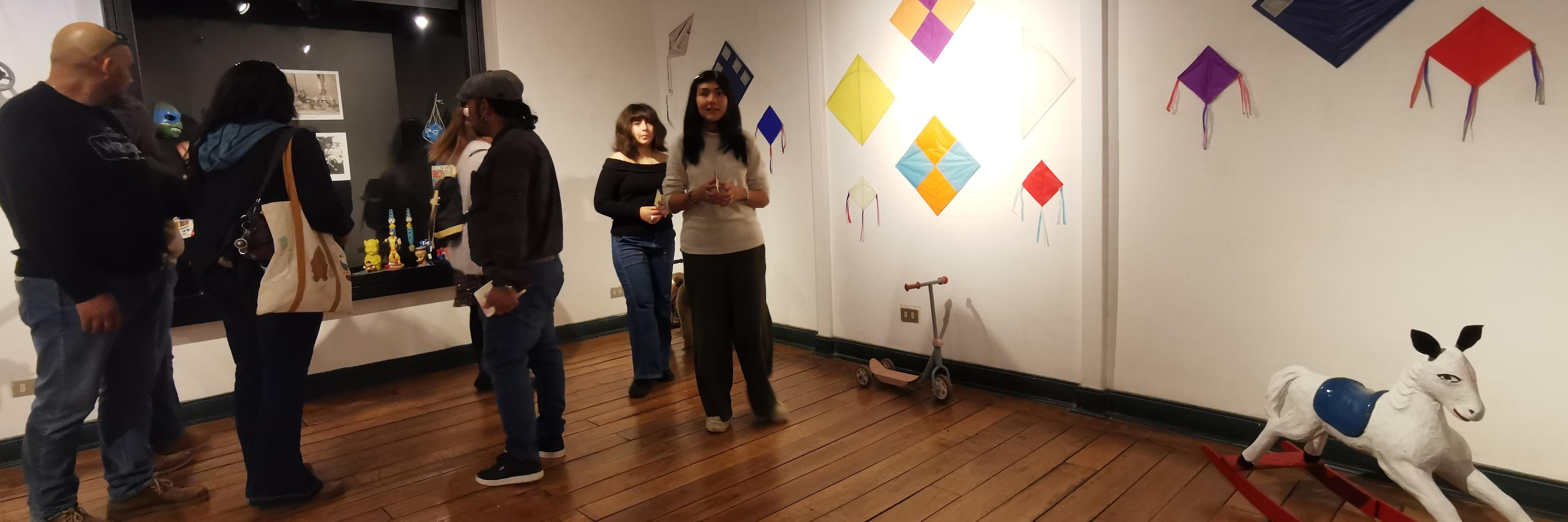 sala de exposición con personas, en la pared blanca se observan volantines de colores