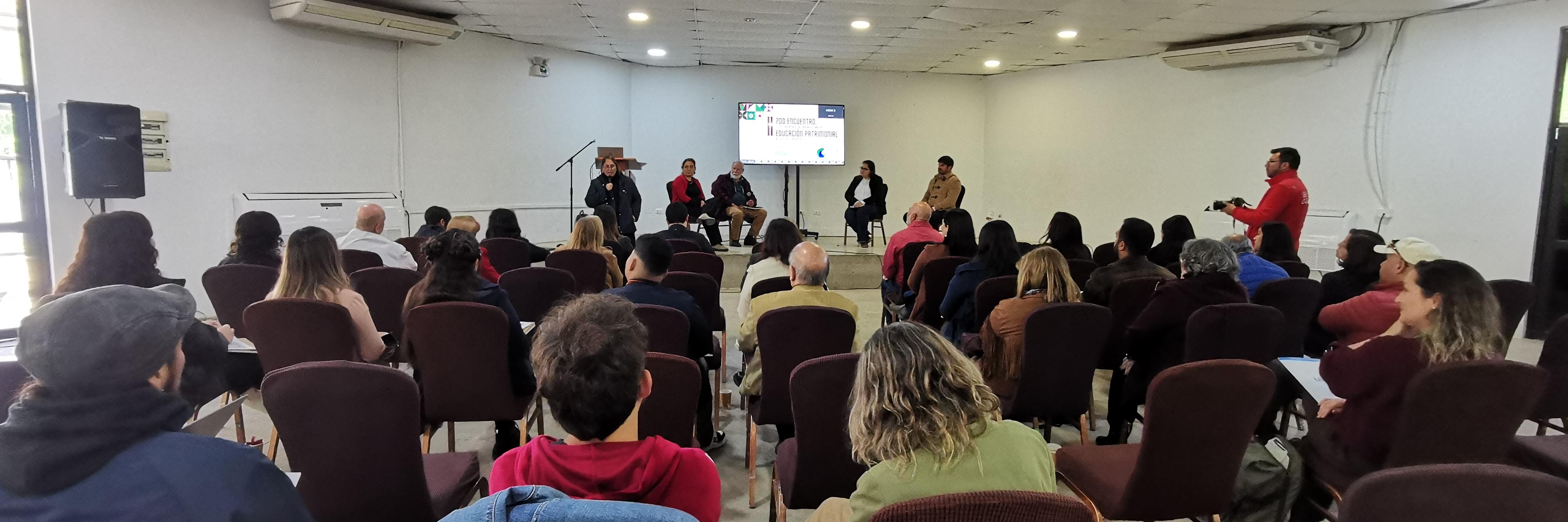 personas sentadas en un salón escuchan a los expositores que presentan en el escenario