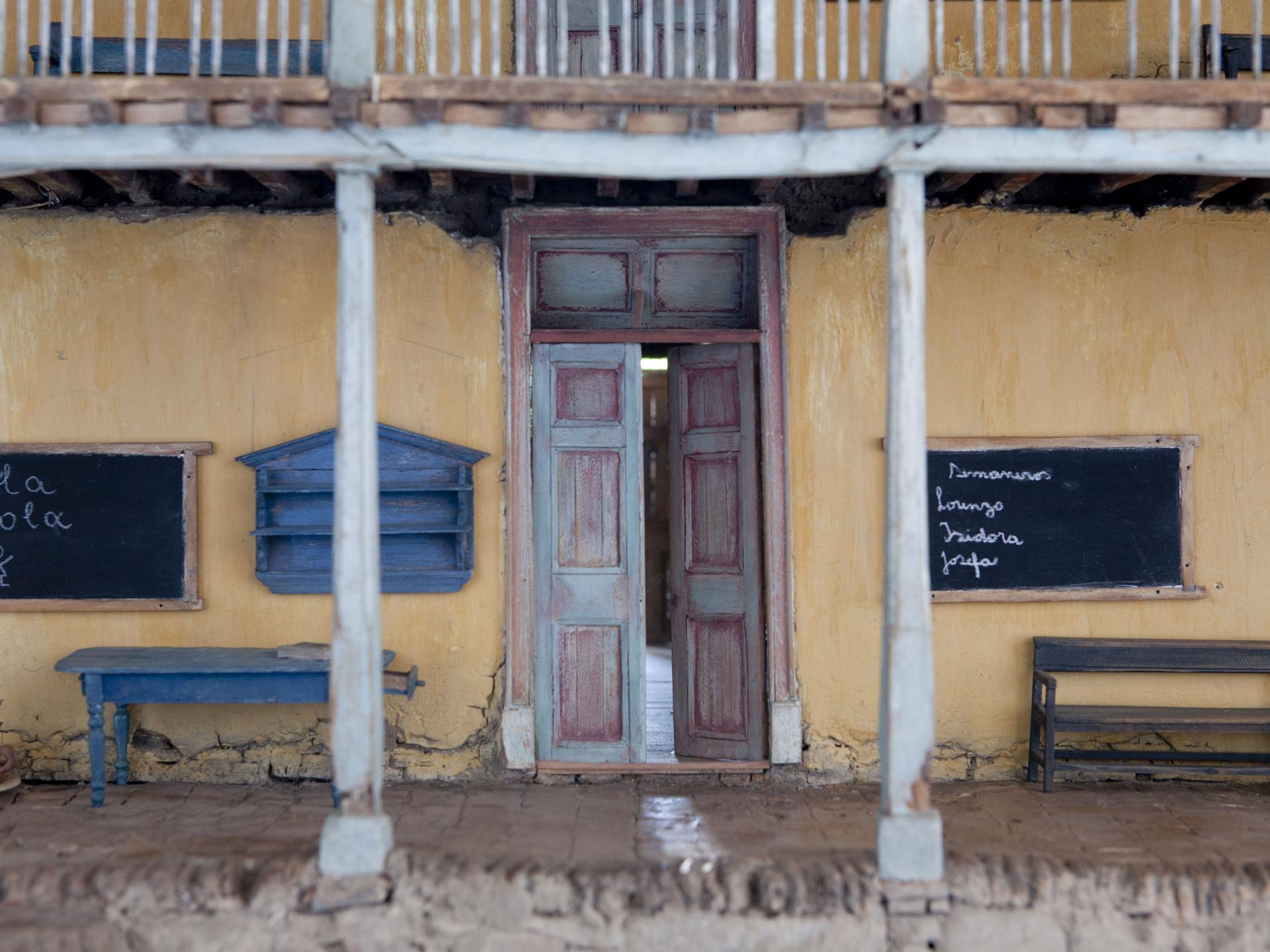 Maqueta de una vivienda tradicional. Se muestra una entrada de un inmueble de dos niveles, se identifica una fachada continua en tono beage, en el lugar hay una puerta, dos pizarras de tiza, una mesa azul y una banca de madera. 