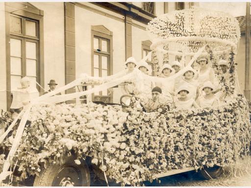 Fotografía monocromática antigua en la que se aprecia un carro alegórico adornado de flores y cintas, en su interior un grupo aproximado de 10 jóvenes disfrazadas para la ocasión. La fotografía registra la fiesta de la primavera en la década de 1920.