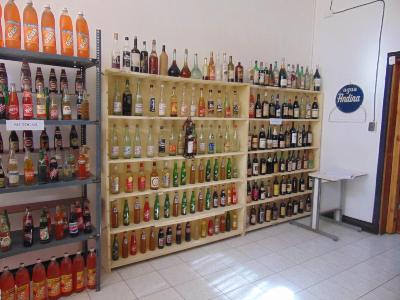 Fotografía en que se aprecia una repisa de varios niveles pegada a la pared, la cual contiene variedad de botellas de licores, bebidas y otros bebestibles de antaño.