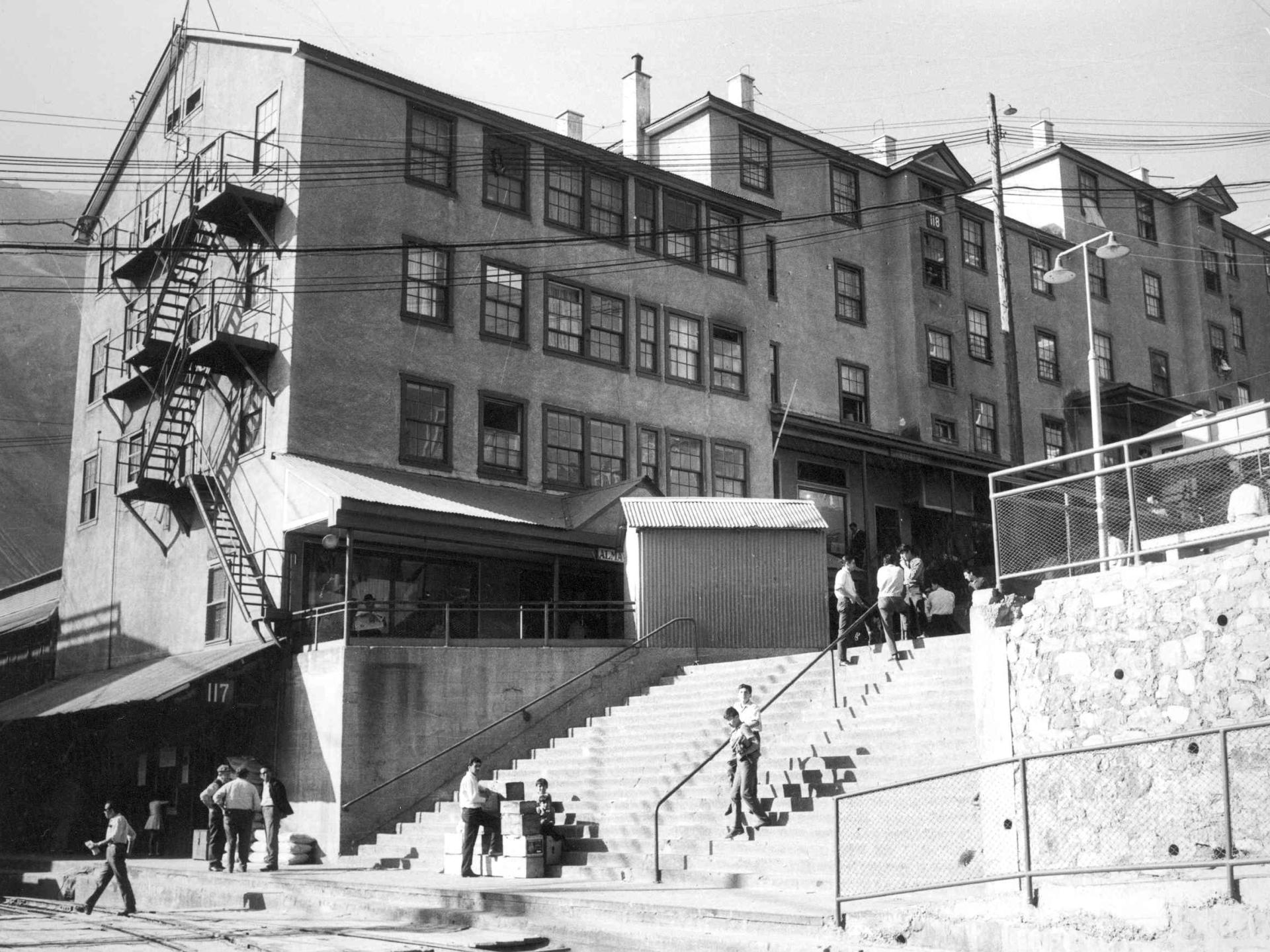 Fotografía monocromática en que se observan en primer plano personas usando la escalera central de Sewell, en segundo plano tres edificios denominados camarotes, lugar en que pernoctaban los mineros. 