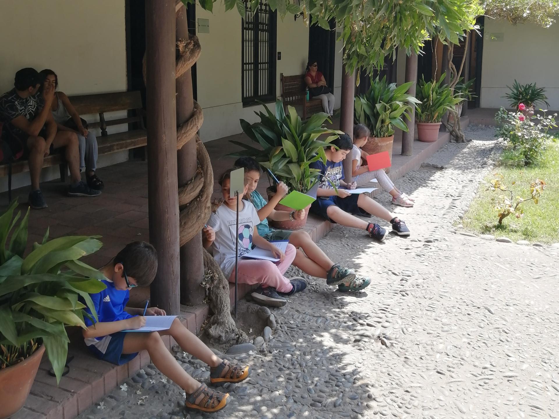 Se aprecia cinco niños y niñas sentadas en el corredor de una casa colonial, escribiendo o dibujando en una libreta. 