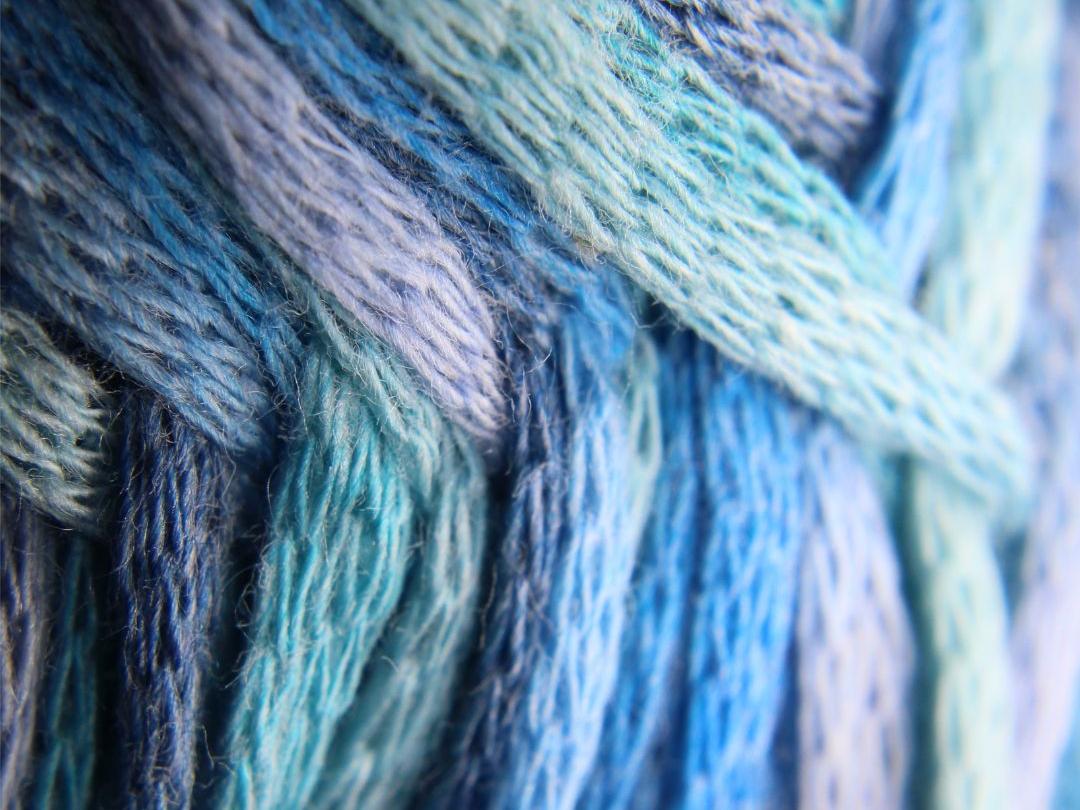 En la imagen se aprecia el detalle de hilos de lana en tonos azul, celeste y purpuras, los que se sobrepone uno sobre otro formando un tejido. 