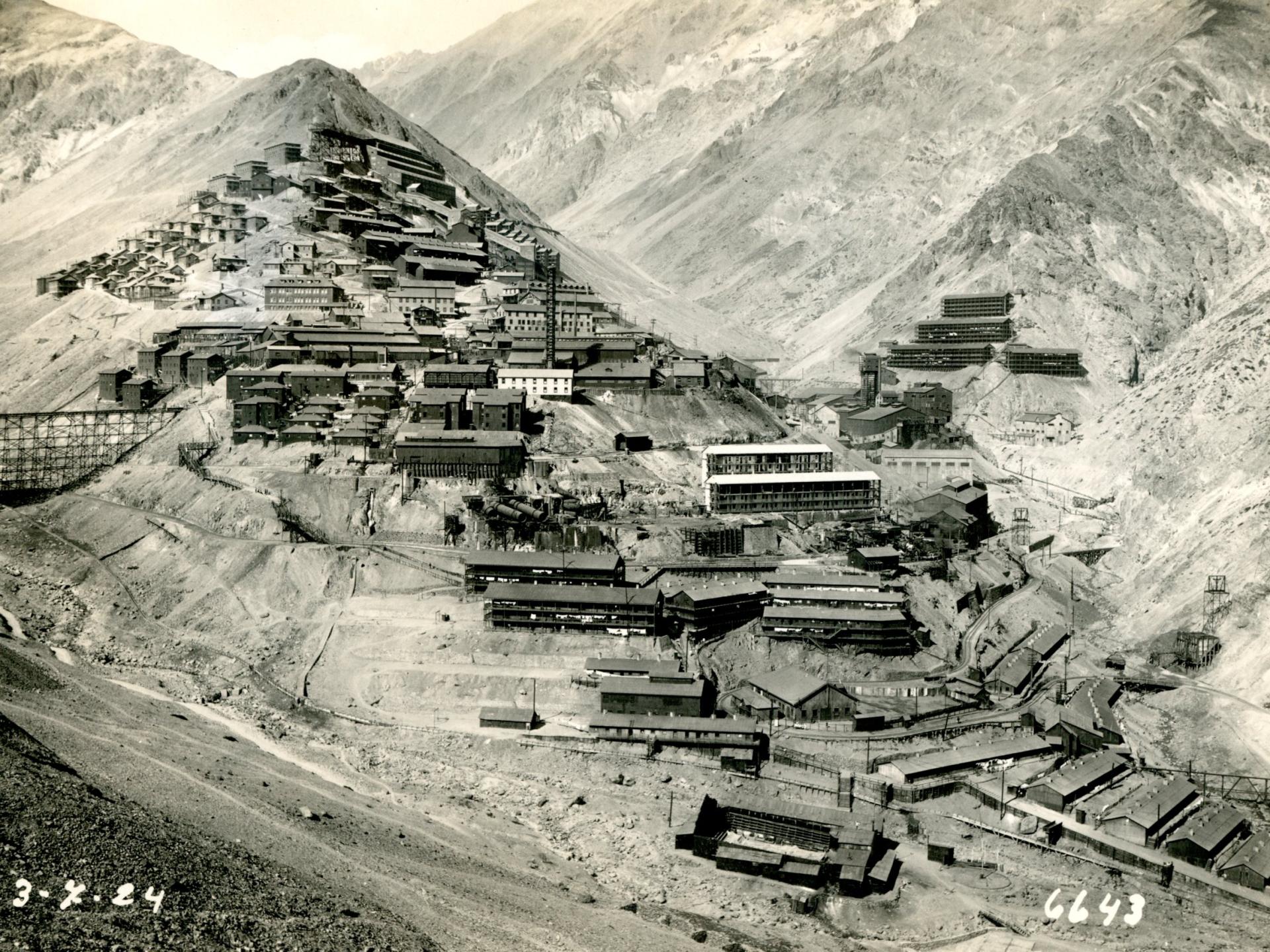 Fotografía en tono sepia del campamento minero Sewell, en ella se aprecian diversos edificios asentados en las montañas de la Cordillera de Los Andes; esta distribución escalonada se genera por su emplazamiento respetando los desniveles del terreno.  