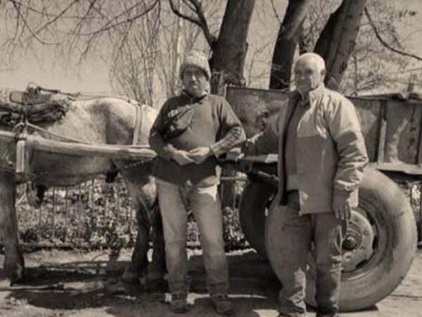Fotografía sepia en que se aprecia una carreta de tracción animal, frente a ella hay dos hombres de frente mirando a la cámara. 