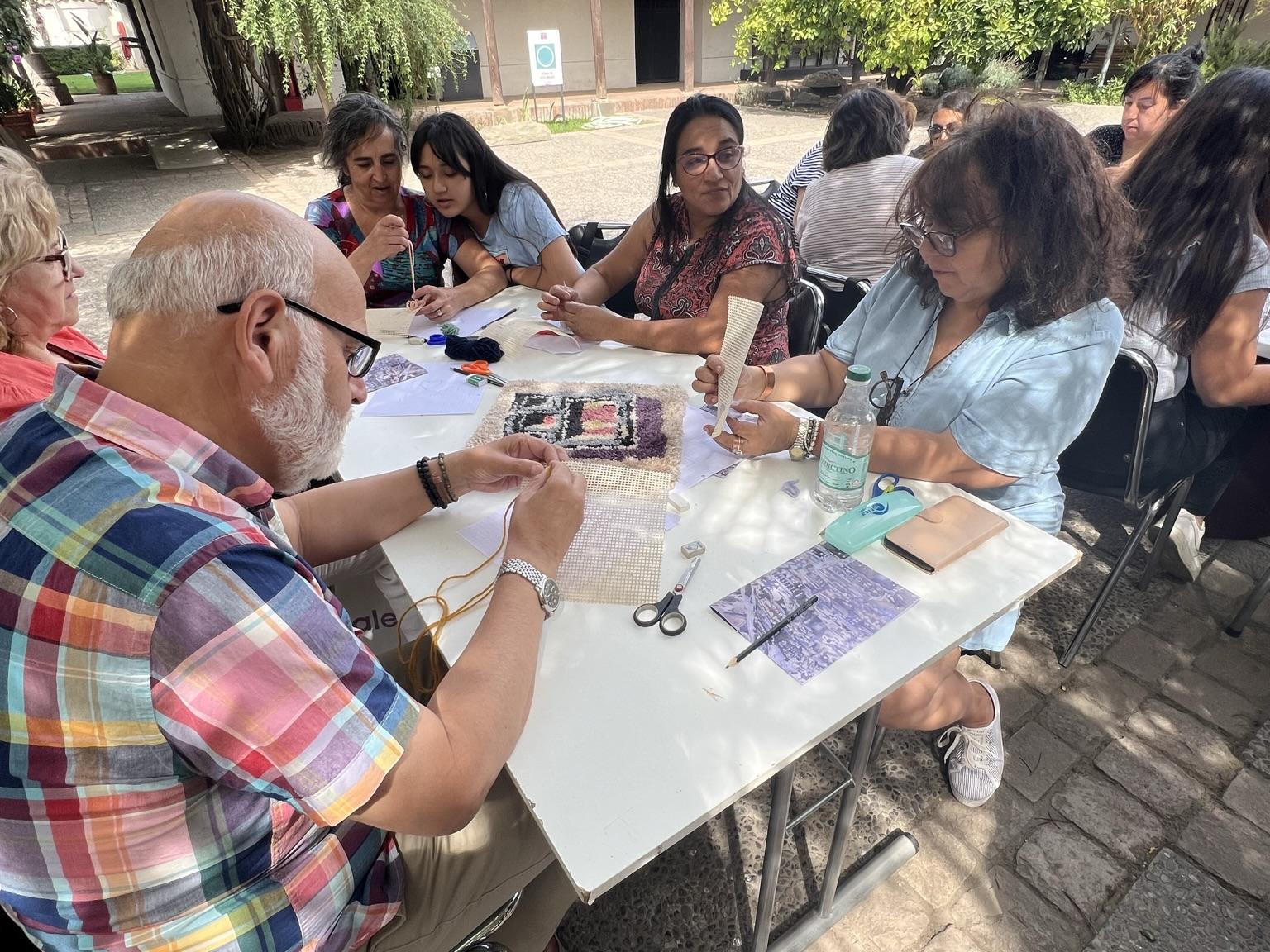 Se aprecian personas adultas sentadas alrededor de una mesa, ellos se encuentran concentrados haciendo un tejido. De fondo, aparece arbustos y la casa colonial que alberga el museo. 