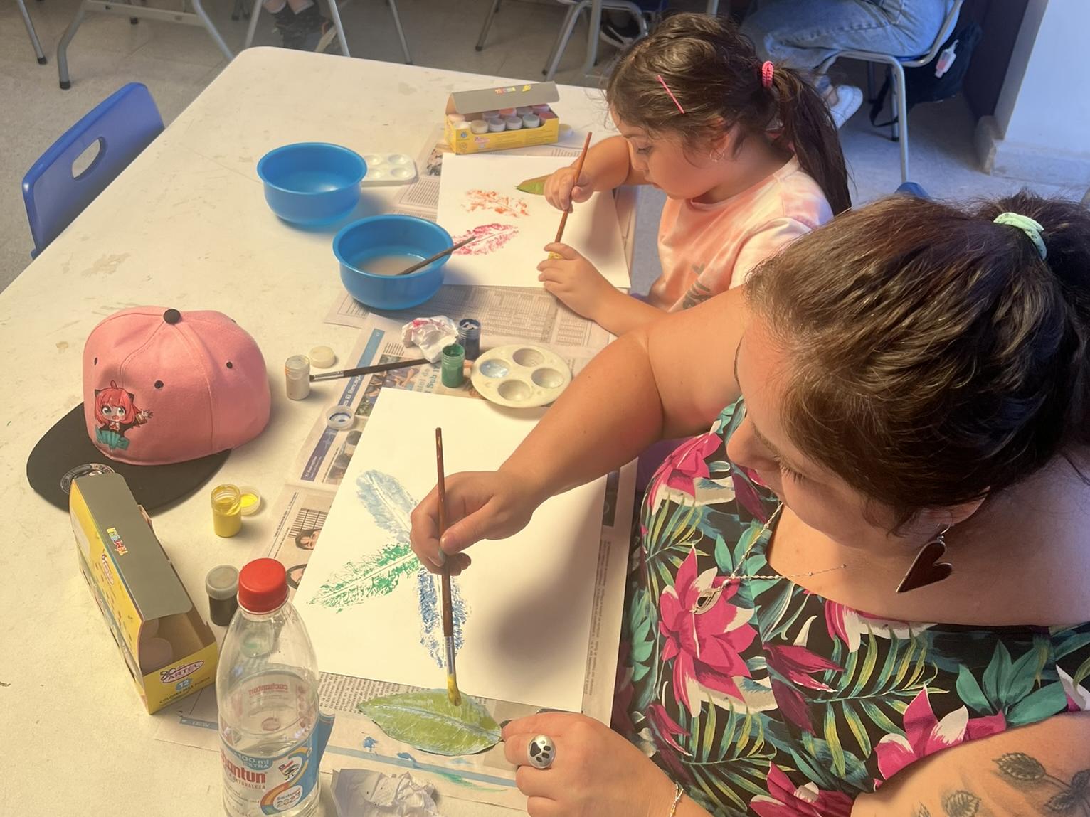 Fotografía en primer plano compuesta por una madre y su hija sentadas pintando con tempera, las pinturas bosquejan estampados de hojas de árboles que van formando nuevas figuras. 