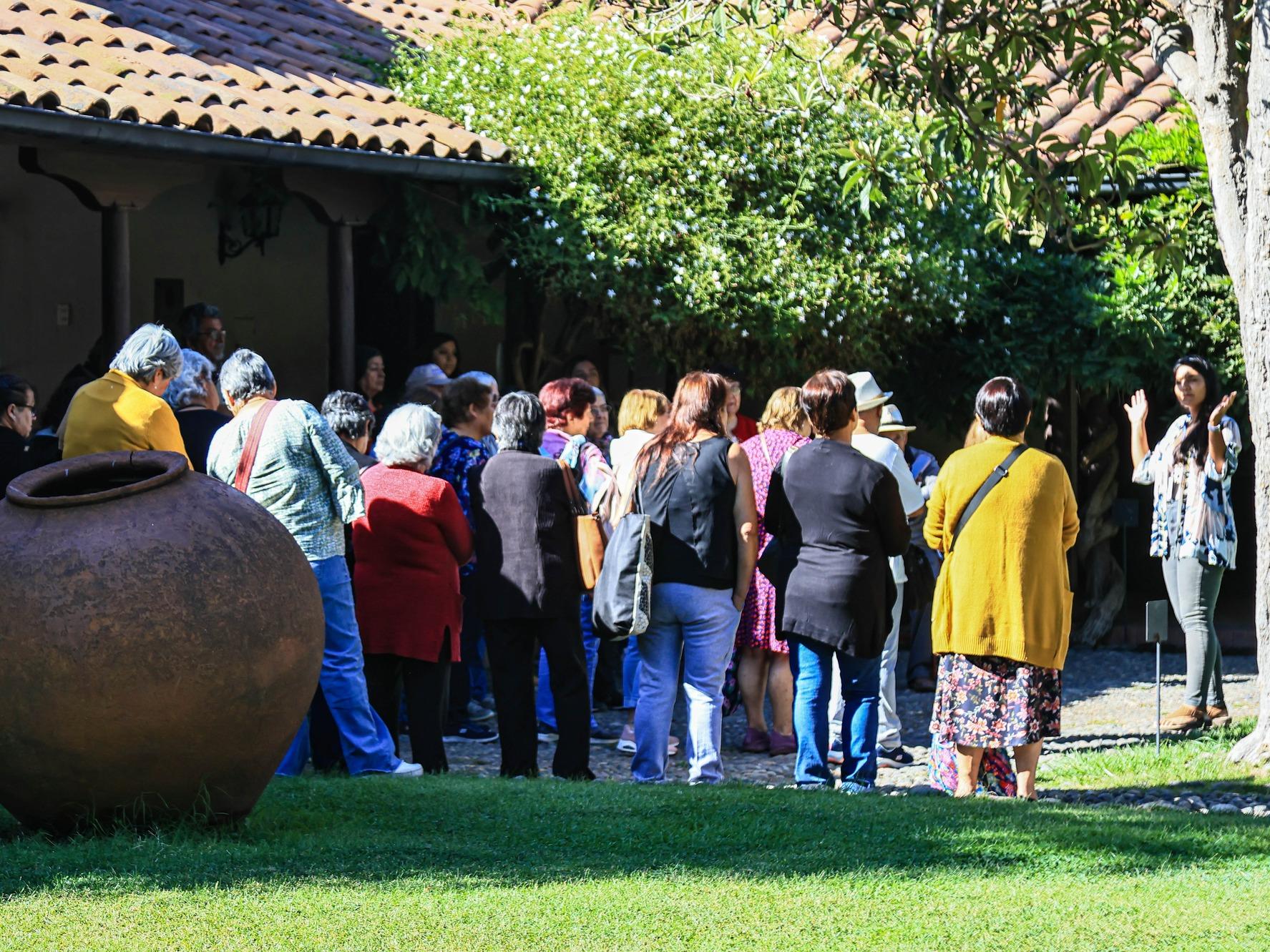 Fotografía panorámica en que se aprecia un grupo de adultos mayores de espalda a la cámara, rodeados de vegetación como un árbol de níspero, un jazmín español y césped. En primer plano se ubica una tinaja de greda (jarrón grande) completando la escena.