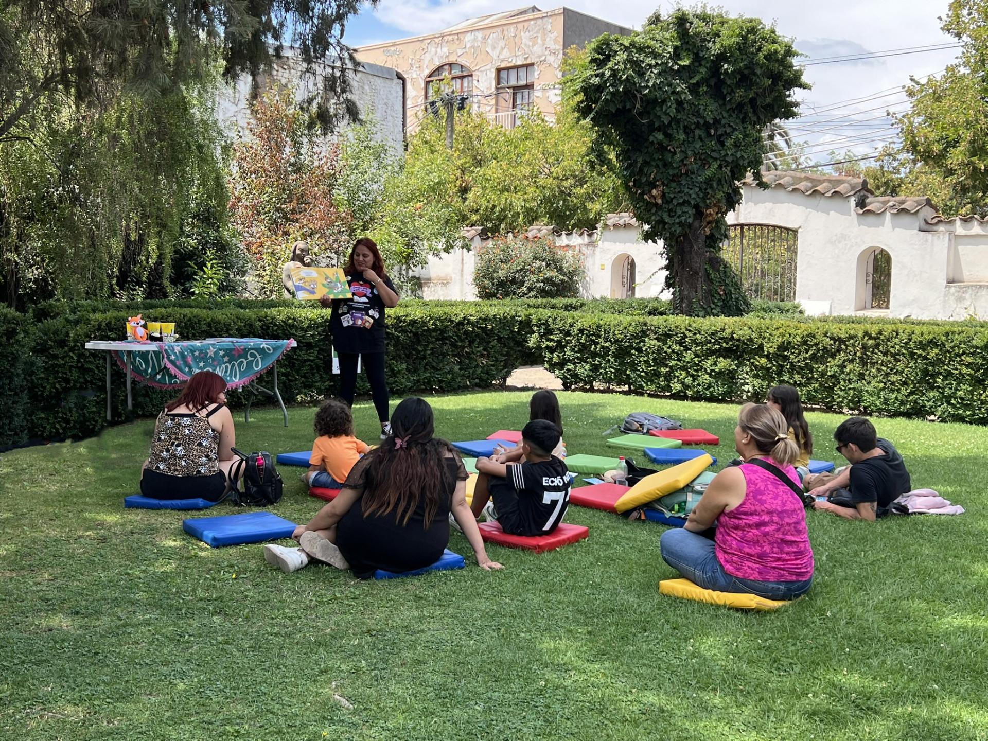 Fotografía panorámica del patio del museo, en el centro hay un grupo de niños y adultos sentados en cojines de colores dispuestos en el césped. De pie, una mujer que es la mediadora de la actividad, ella señala un libro abierto que tiene en su mano. En el fondo de la imagen algunos arboles altos. 