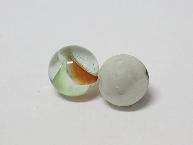 Imagen de fondo gris, en el centra de aprecian dos canicas o bolitas. La primera es de vidrio y en su interior se ven unas líneas verdes y naranja. A su costado hay otra bolita de color blanco.