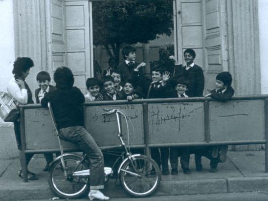 Fotografía monocromatica del ingreso de una escuela urbana, en el centro iun grupo de niños de distintas edades apoyados en una barrera metalica.
