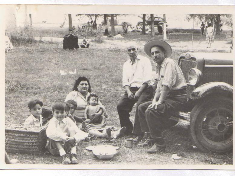 Fotografía monocromática, compuesta de una escena familiar de 7 integrantes que disfrutan un día de picnic.Sentados en el capo de un automóvil, un anciano y un hombre adulto. En el suelo, sentado en el cesped tres niños de edades similares, una mujer con un bebé en sus brazos.