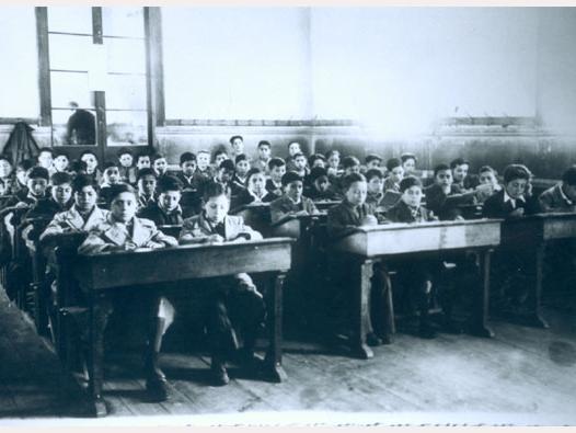 Fotografía monocromatica de una sala de clases en el Instituto O'Higgins de Rancagua. En ella se aprecia un grupo de niños sentados en sus pupitres dobles, la sala está distribuida en tres filas, en el fondo se observa una pared con una ventana de gran tamaño que aparentemente permite mirar hacia otra sala de clases.