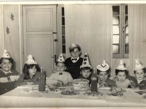 Fotografía monocromática compuesta de una celebración de cumpleaños infantil, sentados a la mesa 7 niños y niñas con gorrito de papel, miran a la cámara. En la mesa hay una torta de crema, bebidas y platos con golosinas diversas.