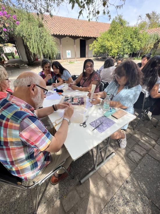 Se aprecian personas adultas sentadas alrededor de una mesa, ellos se encuentran concentrados haciendo un tejido. De fondo, aparecen arbustos y la casa colonial que alberga el museo. 