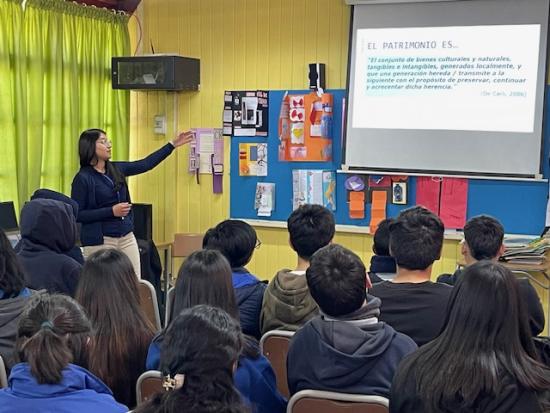 Fotografía de charla, en primer plano distintos estudiantes sentados, al fondo de la imagen se aprecia una mujer señalando un panel que tiene escrita la frase “El Patrimonio es…”