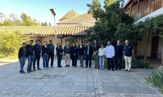 Fotografía en que se aprecian 15 personas adultas en fila, posando hacia la foto. De fondo hay algunos árboles y una planta de una casa colonial, murallas blancas de adobe y techos de teja en tonos marrón. 