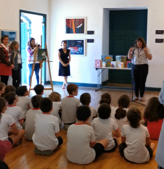Niños y niñas sentados en el piso escuchan a una persona de pied delante que les habla. El lugar es un salon donde hay cuadros colagdos en las paredes. 