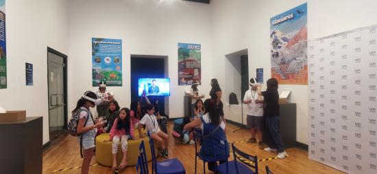 Niñas y niños de pie y sentados en un salon. Una niña con lentes VR en la cabez y las otras niñas la observan.