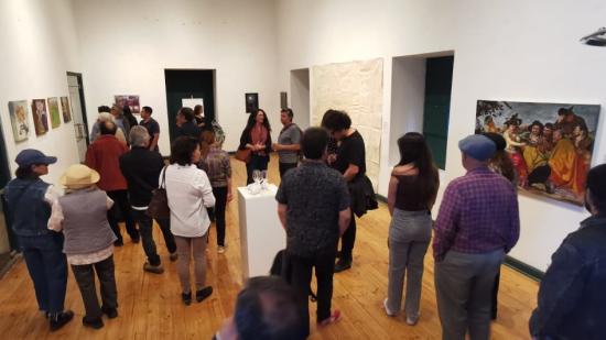 En un salón de paredes blancas y piso de madera mujeres y hombres conversan entre sí. en ls paredes se observan cuadros y tambien una escultura en un pedestal.