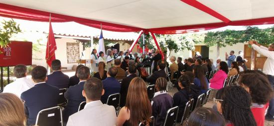 personas sentadas bajo un carpa de color blanco, al fondo personas en un coro de pie