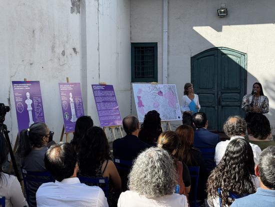 En en espacio exterior de un edificio con muros blancos y una puerta de madera, Dos mujeres están de pie, una de ellas habla con un micófono a las personas que escuchan sentadas