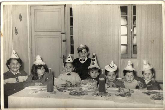 Fotografía monocromática compuesta de una celebración de cumpleaños infantil, sentados a la mesa 7 niños y niñas con gorrito de papel, miran a la cámara. En la mesa hay una torta de crema, bebidas y platos con golosinas diversas. 